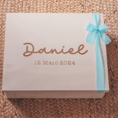 Caixa branca com laço azul claro e texto dourado em superfície de ráfia