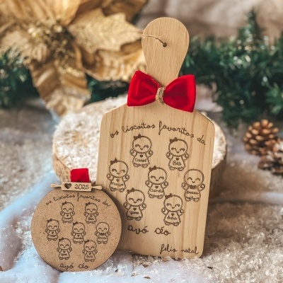 Tábua de madeira decorada com laço vermelho e figura de bolachas desenhadas com nomes, placa redonda com texto semelhante; cenário natalício com neve artificial e pinhas.