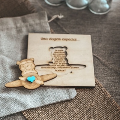 Convite de madeira com desenho de ursinho em avião e coração azul sobre tecidos rústicos