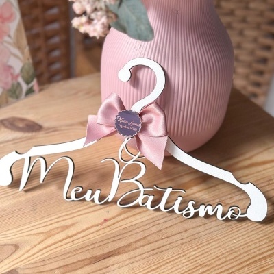 Cabide branco com texto Meu Batismo e laço cor-de-rosa, em cima de mesa de madeira, junto a vaso rosa com flores.
