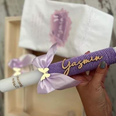 Vela branca decorada com fita lilás, laço com borboleta dourada e nome Yasmin em malha roxa num ambiente rústico