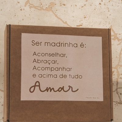 Caixa de cartão castanha com etiqueta branca e texto em português sobre ser madrinha
