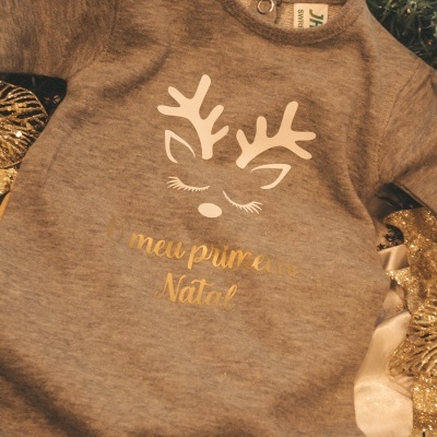 Macacão cinzento de bebé com desenho de rena e texto 'O meu primeiro Natal' rodeado de decoração natalícia