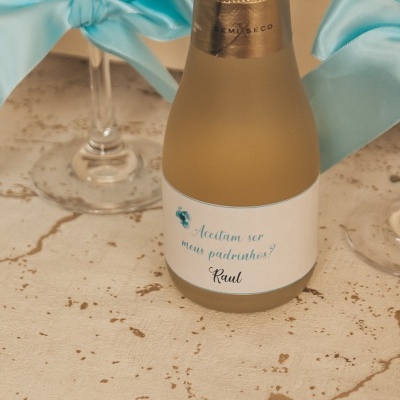 Pequeno frasco de vidro fosco com rótulo branco entre copos com laços azuis