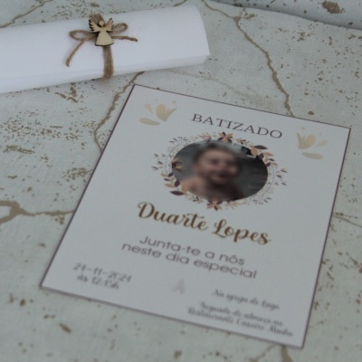 convite branco de batizado com decoração floral pastel, texto e foto circular desenfocada perto de envelope enrolado com fita e anjo