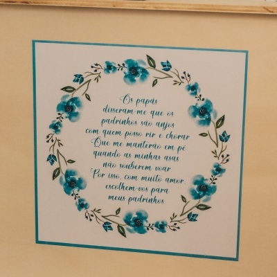 Quadro com moldura de madeira clara com uma coroa de flores azuis e texto em português no centro.