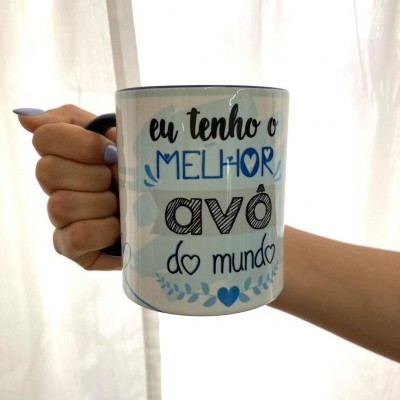 Caneca branca com texto 'eu tenho o MELHOR avô do mundo' segurada por mão com unhas azul claro