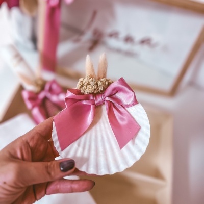 Concha branca com laço rosa e flores secas decorativas, segurada por mão com unhas roxas
