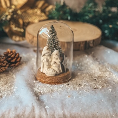 Miniatura de presépio branco numa base de cortiça com globo transparente, em ambiente natalício.