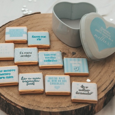 Caixa metálica em forma de coração com chocolates quadrados com mensagens em português, sobre base de madeira redonda