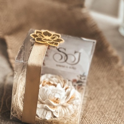 Caixa transparente com flor branca, fita dourada e etiqueta SOJ