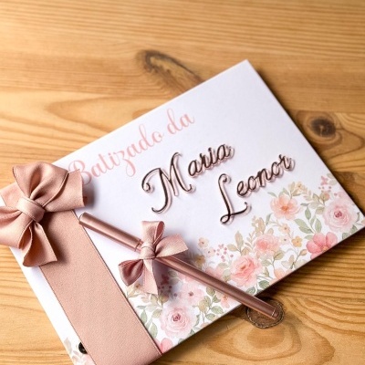 Livro de assinaturas decorado para batizado com flores, fita rosa e caneta rosa sobre mesa de madeira.