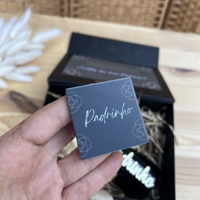 Placa em acrílico cinza escuro com texto branco 'Padrinho' segurada à mão com caixa preta e palha decorativa ao fundo.