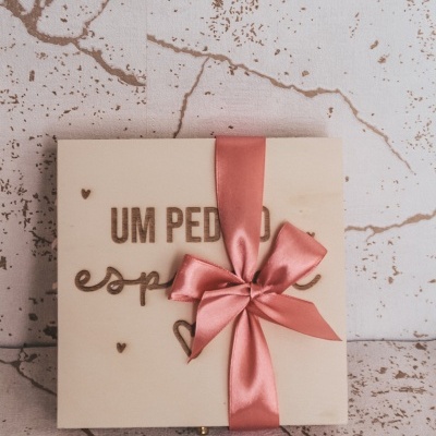 Caixa de madeira clara com laço rosa e texto gravado, sobre mesa com padrão, parede clara com manchas castanhas.