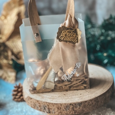Saco translúcido com alça castanha, laço e placa dourada 'Feliz Natal' com utensílios dourados dentro sobre base de madeira