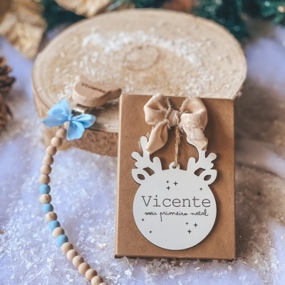 Etiqueta natal decoração em forma de rena branca com texto Vicente meu primeiro natal e corrente de contas de madeira e azul claro sobre neve artificial