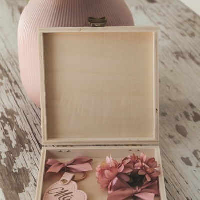 Caixa de madeira aberta com coração rosa escrito 'Mãe' e coroa de flores rosa ao lado, sobre mesa rústica com vaso rosa ao fundo.