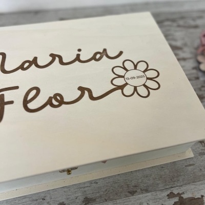 Caixa de madeira clara com gravação de 'Maria Flor' e data numa flor, sobre madeira rústica.