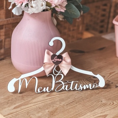 Cabide branco decorativo com texto Meu Batismo, laço rosa e coração com nome Luna ao lado de vaso rosa com flores artificiais