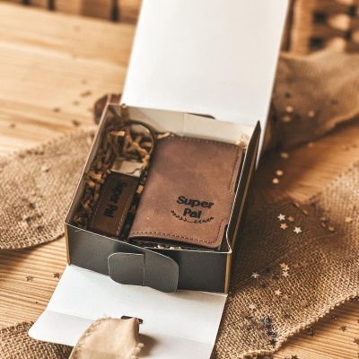 Conjunto de carteira e porta-chaves em pele castanha dentro de caixa preta aberta sobre superfície de madeira com tecido rústico e estrelas decorativas.