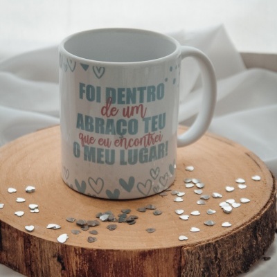 Caneca branca com texto colorido sobre base de madeira com corações prateados, fundo de tecido branco.