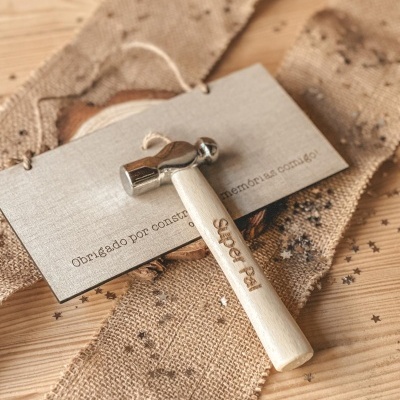 Martelo pequeno em madeira clara com cabeça prateada e texto gravado no cabo sobre cartão cinza e tecido bege