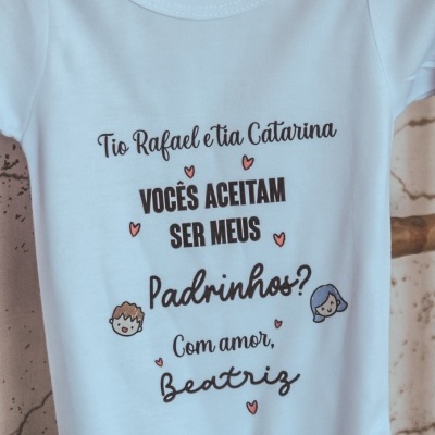 Body branco de bebé com texto em português e pequenos desenhos coloridos, pendurado num cabide cinzento