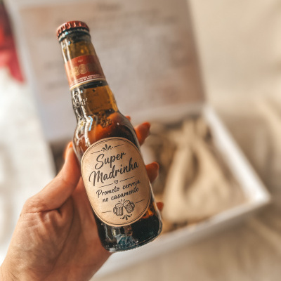 Garrafa de cerveja com rótulo personalizado Super Madrinha numa mão com fundo desfocado