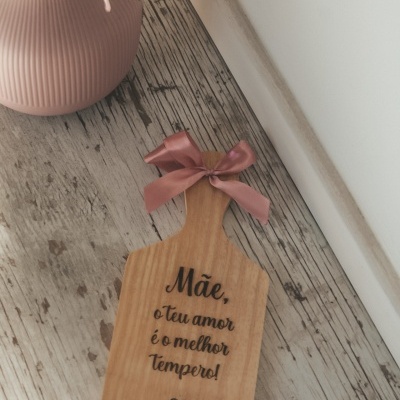 Tábua de madeira gravada com texto em português e fita rosa, junto a vaso rosa com flores