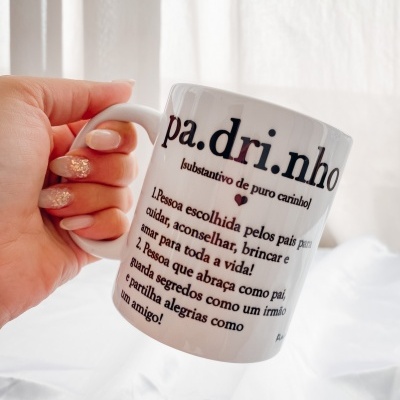 Caneca branca com texto em português sobre padrinho, segurada por mão com unhas decoradas