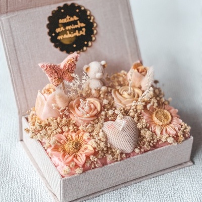 Arranjo floral decorativo com flores de cera, ursinho e coração em caixa de tecido bege com etiqueta preta com texto