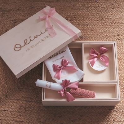 Caixa de presente em madeira clara com acessórios decorados com laços rosa sobre tapete castanho.