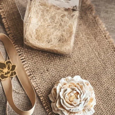 Flor decorativa branca com detalhes dourados e fita bege sobre tecido de juta
