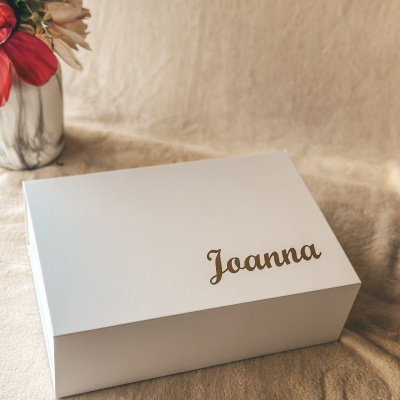 Caixa branca com o nome Joanna em letras douradas ao lado de um vaso com flores