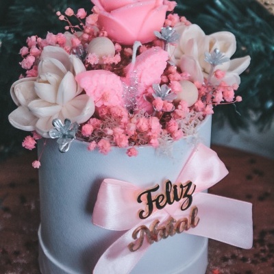 Arranjo decorativo de Natal com vela rosa e flores em recipiente branco com laço e frase Feliz Natal