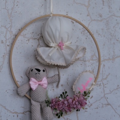 Decoração pendurada com ursinho de crochê cinza, gravata rosa, chapéu branco com cruz rosa e flores cor-de-rosa