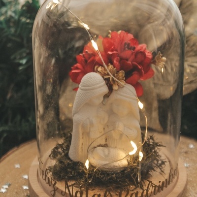 Presépio branco sob cúpula de vidro com flores vermelhas e luzes, base de madeira com texto festivo.
