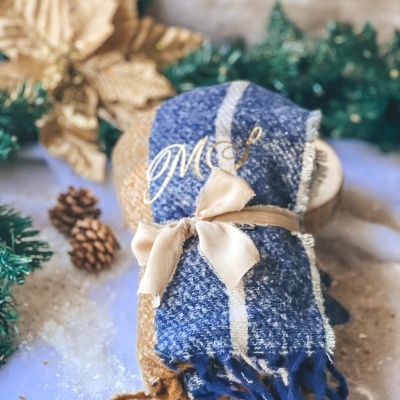Conjunto de mantas de lã em castanho e azul com fita e iniciais douradas, decoradas com elementos naturais em fundo claro.