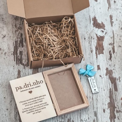 Caixa de presente aberta com enchimento de palha, texto 'Santiago 19-06-2023', caixa pequena de madeira com texto 'pa.dri.nho' e laço azul sobre madeira envelhecida