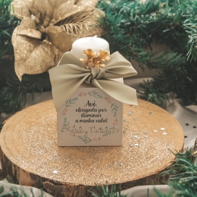 Caixa de presente branca decorada com laço verde-acinzentado e flor dourada sobre base de madeira com folhas douradas e verdes