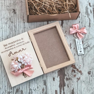 Moldura de madeira clara com cartão decorativo, flores rosa e laço, caixa de papelão com palha, laço rosa com etiqueta, fundo madeira desgastada