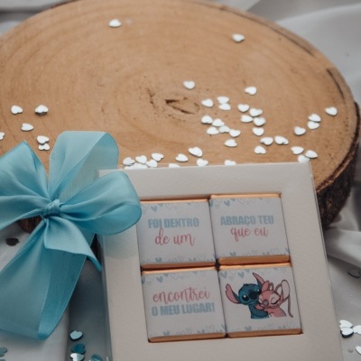 Caixa branca com quatro chocolates com textos em português e personagens Stitch e Angel, laço azul, sobre tecido branco e base de madeira com corações.