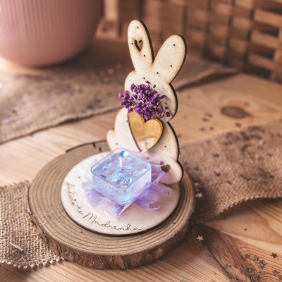 Suporte decorativo de madeira em forma de coelho com flores secas e recipiente lilás sobre base redonda de madeira.
