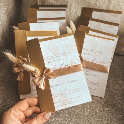 Convites de batizado em papel branco texturado com envelope dourado e decoração com fita castanha e flores secas.