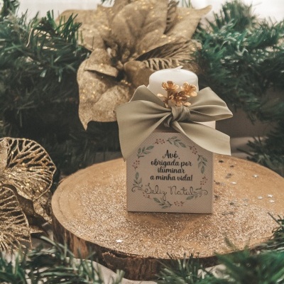 Vela decorativa branca com fita verde e caixa com mensagem de Natal em fundo de folhagem e flores douradas