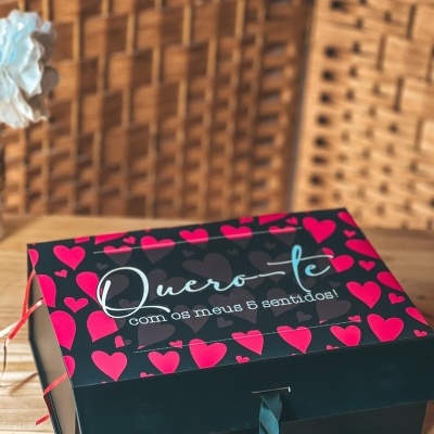 Caixa de presente preta com corações vermelhos e texto Quero-te com os meus 5 sentidos! numa mesa de madeira.