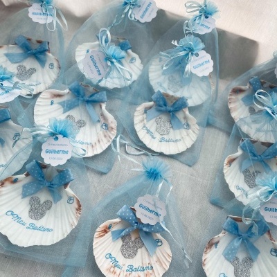 Conchas decorativas brancas com texto azul e laços azuis dentro de saquinhos azuis translúcidos.