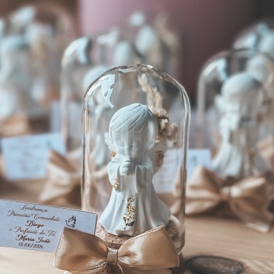 Figurinha de menino rezando em cúpula de vidro com laço dourado e etiqueta de lembrança de comunhão.