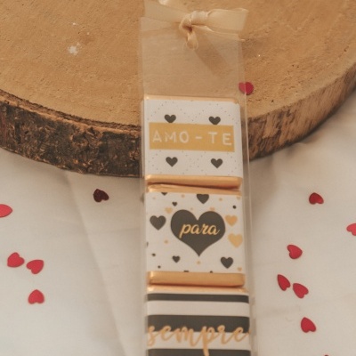 Conjunto de três barras de chocolate com mensagens em embalagem transparente e fita de laço bege sobre mesa branca com corações vermelhos de papel.