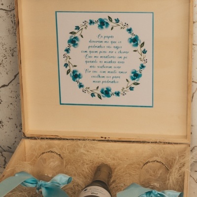 Caixa de madeira com garrafa de champanhe, copos com laços azuis e cartaz decorativo com texto e flores azuis